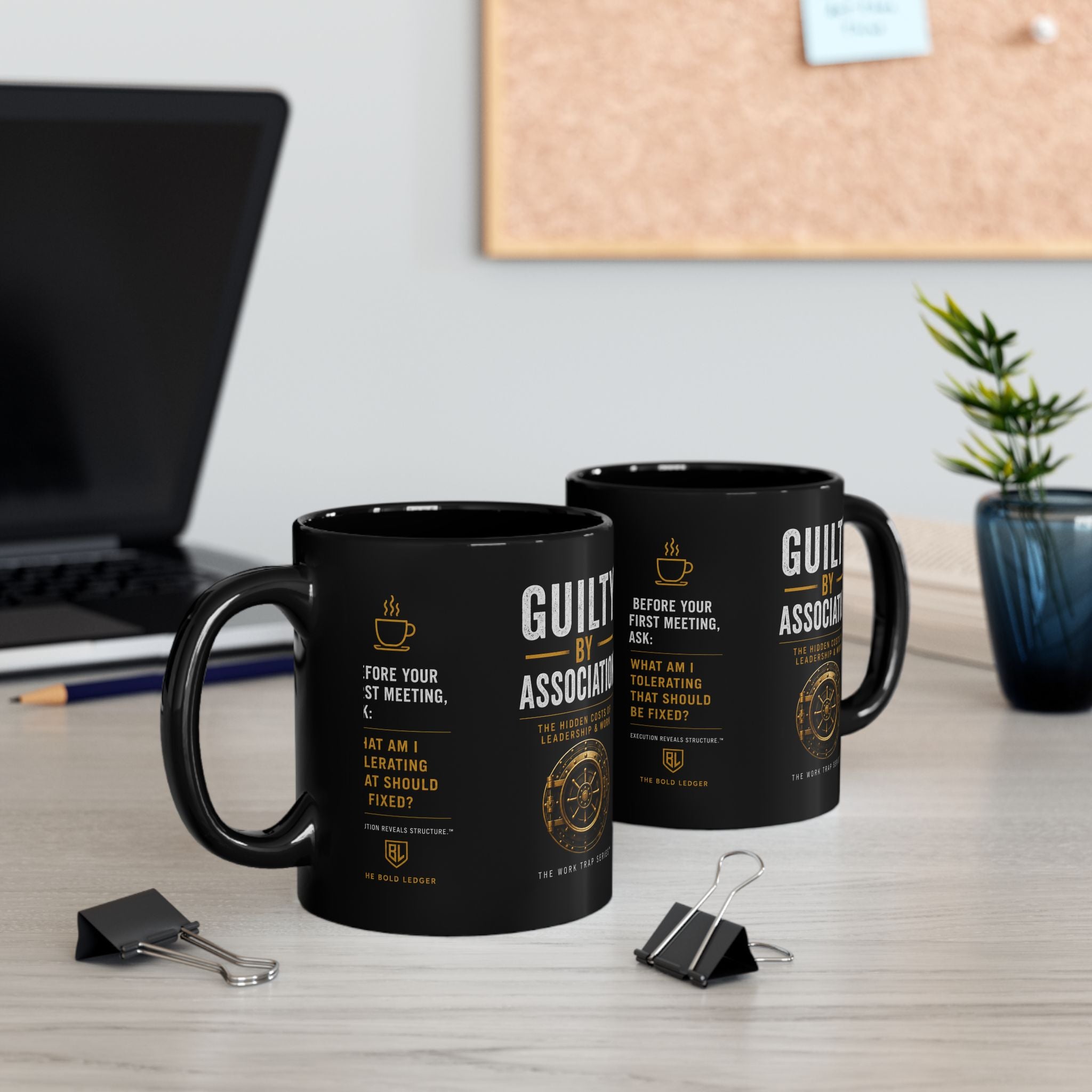 Two black mugs with text on a desk with a laptop and plant in the background